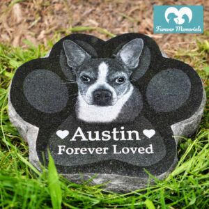 Black granite paw-shaped pet memorial stone engraved with a portrait of Austin the dog and the inscription “Forever Loved,” placed in grass in a garden setting.