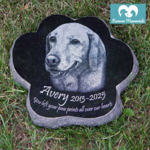 Custom engraved black granite paw-shaped pet memorial for dog Avery with detailed portrait engraving and inscription, placed on grass in an outdoor garden setting