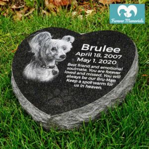 Black granite heart-shaped pet memorial stone engraved with a portrait of Brulee the dog, featuring the dates April 18, 2007 – May 1, 2020, placed in grass in a garden setting.