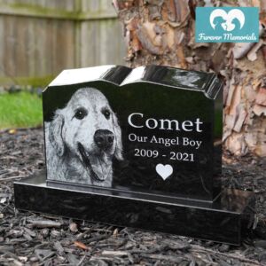Black granite book-style pet memorial engraved with a portrait of Comet the dog and the inscription “Comet – Our Angel Boy – 2009–2021,” displayed in a garden setting.