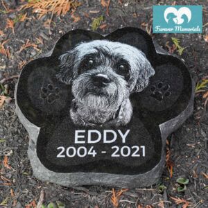 Black granite paw-shaped pet memorial engraved with a portrait of Eddy the dog and the inscription “Eddy 2004–2021,” placed in a garden bed.