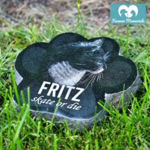 Black granite paw-shaped pet memorial stone engraved with a portrait of Fritz the cat and the inscription “Skate or die,” placed in grass in a garden setting.