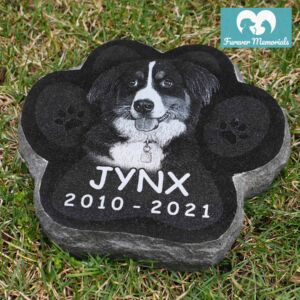 Black granite paw-shaped pet memorial engraved with a portrait of Jynx the dog and the inscription “Jynx 2010–2021,” placed on grass.