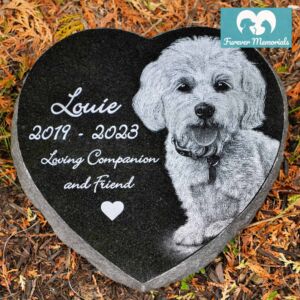 Heart-shaped black granite pet memorial engraved with a portrait of Louie the dog, including the inscription “Louie 2019–2023 Loving Companion and Friend,” placed in a garden bed with autumn leaves.