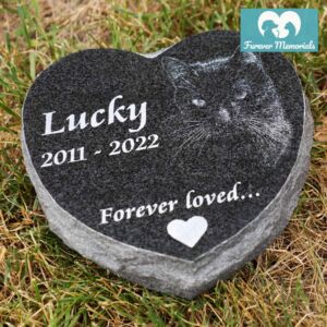 Black granite heart-shaped pet memorial stone engraved with a portrait of Lucky the cat, including the dates 2011–2022 and the inscription “Forever loved…,” placed in grass in a garden setting.
