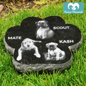Black granite paw-shaped pet memorial stone engraved with portraits of three dogs named Mate, Scout, and Kash, placed in grass in an outdoor garden setting.