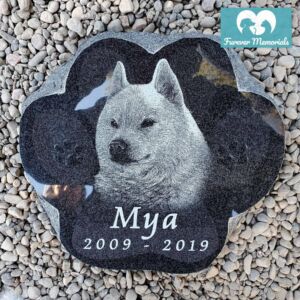 Black granite paw-shaped pet memorial stone engraved with a portrait of Mya the dog and the dates 2009–2019, placed on decorative garden stones.