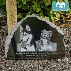 Rustic black granite pet memorial stone engraved with portraits of two dogs named Onyx and Chase, placed in a garden setting with natural stone edges.