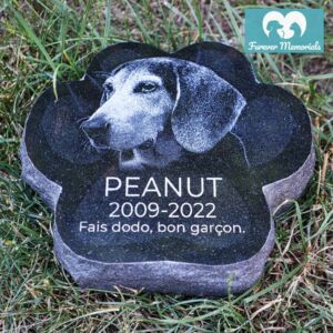 Black granite paw-shaped pet memorial stone engraved with a portrait of Peanut the dog, including the dates 2009–2022 and the inscription “Fais dodo, bon garçon,” placed in grass in a garden setting.