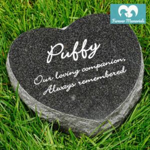 Black granite heart-shaped pet memorial stone engraved with the name Puffy and the inscription “Our loving companion. Always remembered,” placed in grass in a garden setting.