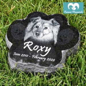 Black granite paw-shaped pet memorial stone engraved with a portrait of Roxy the dog and the dates June 2010 – February 2020, placed in grass in a garden setting.