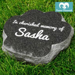Black granite paw-shaped pet memorial stone engraved with the inscription “In cherished memory of Sasha,” placed in grass in a garden setting.