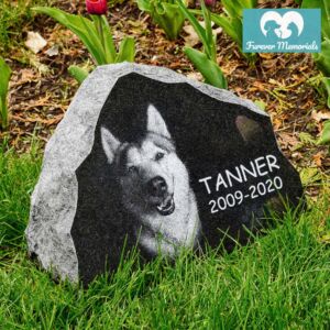 Rustic black granite pet memorial stone engraved with a portrait of Tanner the dog and the dates 2009–2020, placed in grass in a garden setting.