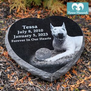 Black granite heart-shaped pet memorial engraved with a portrait of Tessa the Bull Terrier lying beside a ball, personalized with her name, dates (July 8, 2010 – January 5, 2023), and the inscription “Forever In Our Hearts,” placed in a garden setting.