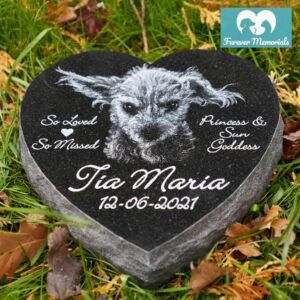 Heart-shaped black granite pet memorial engraved with a portrait of Tia Maria the dog, featuring the inscription “So Loved, So Missed” and the words “Princess & Sun Goddess,” placed in a grassy garden setting.