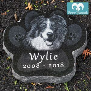 Paw-shaped black granite pet memorial engraved with a portrait of Wylie the dog and the dates 2008–2018, placed in a garden bed.